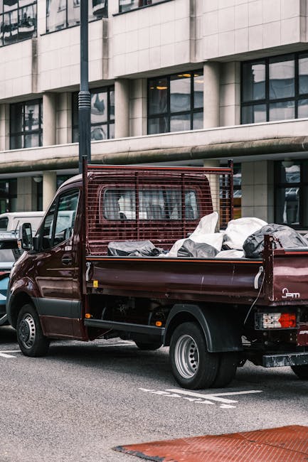A small, dark red flatbed truck parked on an urban street in front of a modern building with large glass windows and a concrete facade. The truck's cargo area is loaded with various types of waste, in