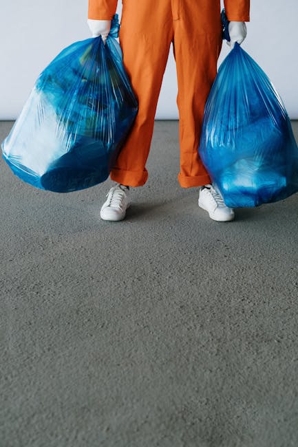 A person standing on a grey carpeted floor, wearing orange pants and white sneakers, holding two large blue plastic rubbish bags filled with waste in each hand. The bags are semi-transparent, revealin